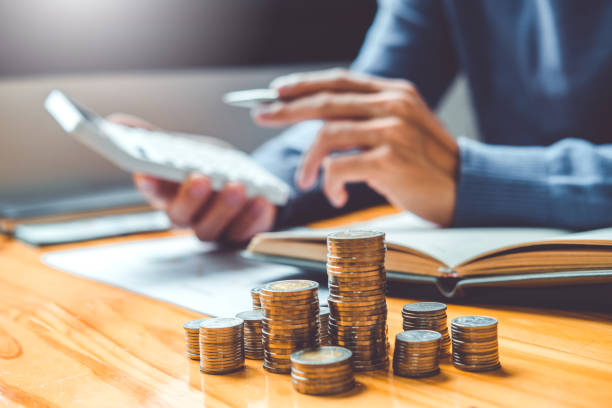Person calculating finances with a calculator and notebook on a desk, next to stacked coins representing savings or investment planning.